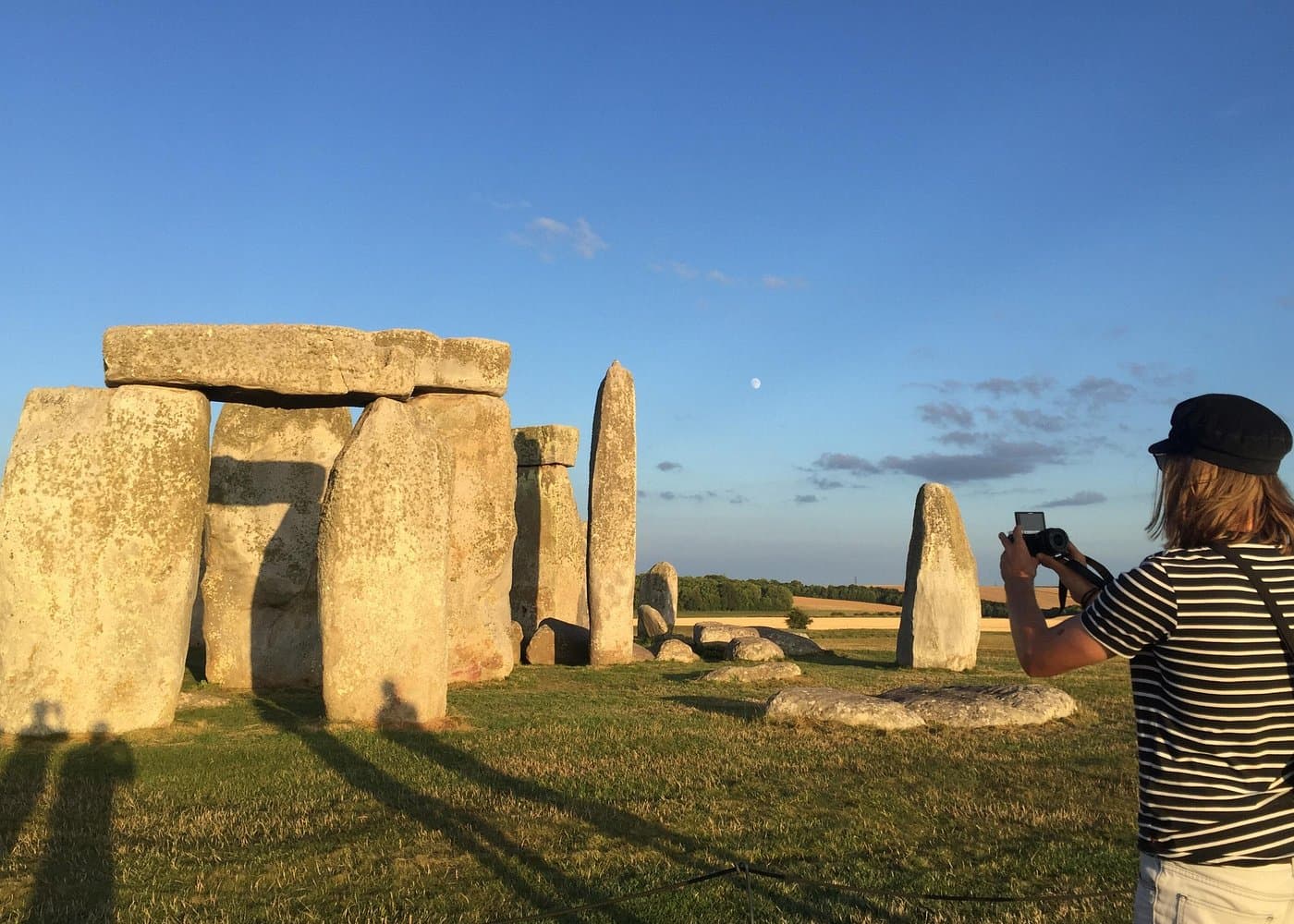 A visitor photographing Stonehenge in warm evening light, with a moon in a clear blue sky.